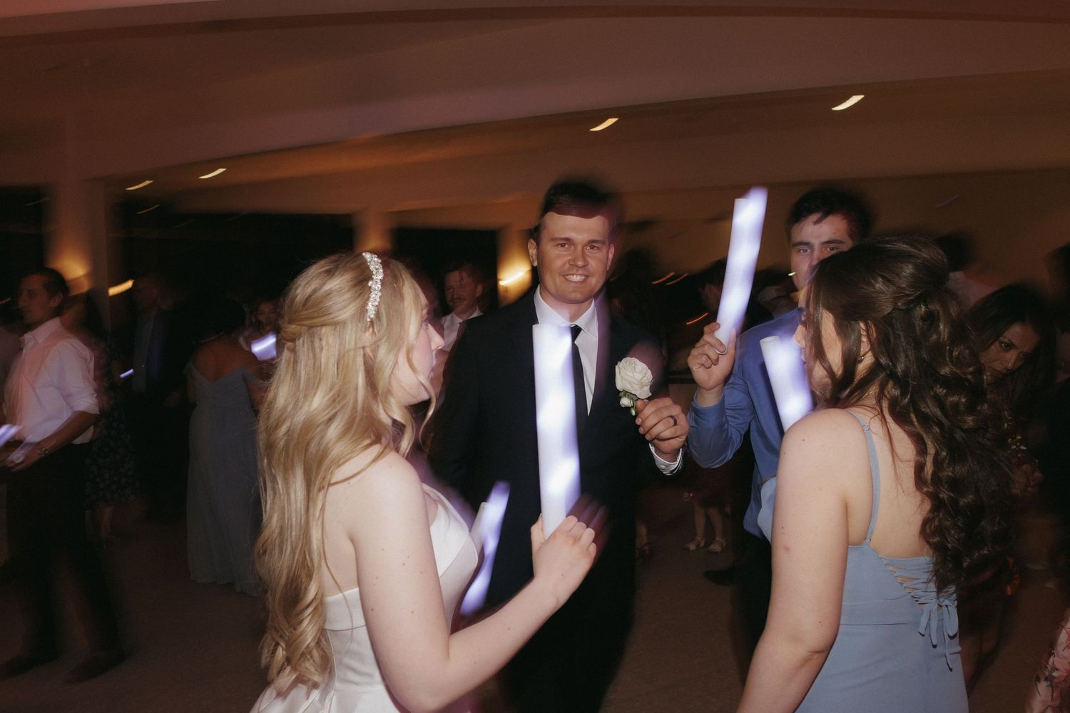 Bride and groom dance joyfully with guests holding glowing light sticks at lively wedding reception party.