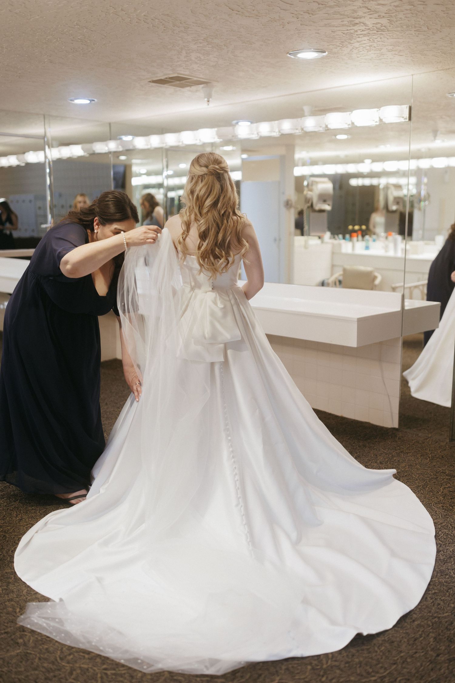Bride in white wedding gown with long train gets veil adjusted by bridesmaid in dressing room.
