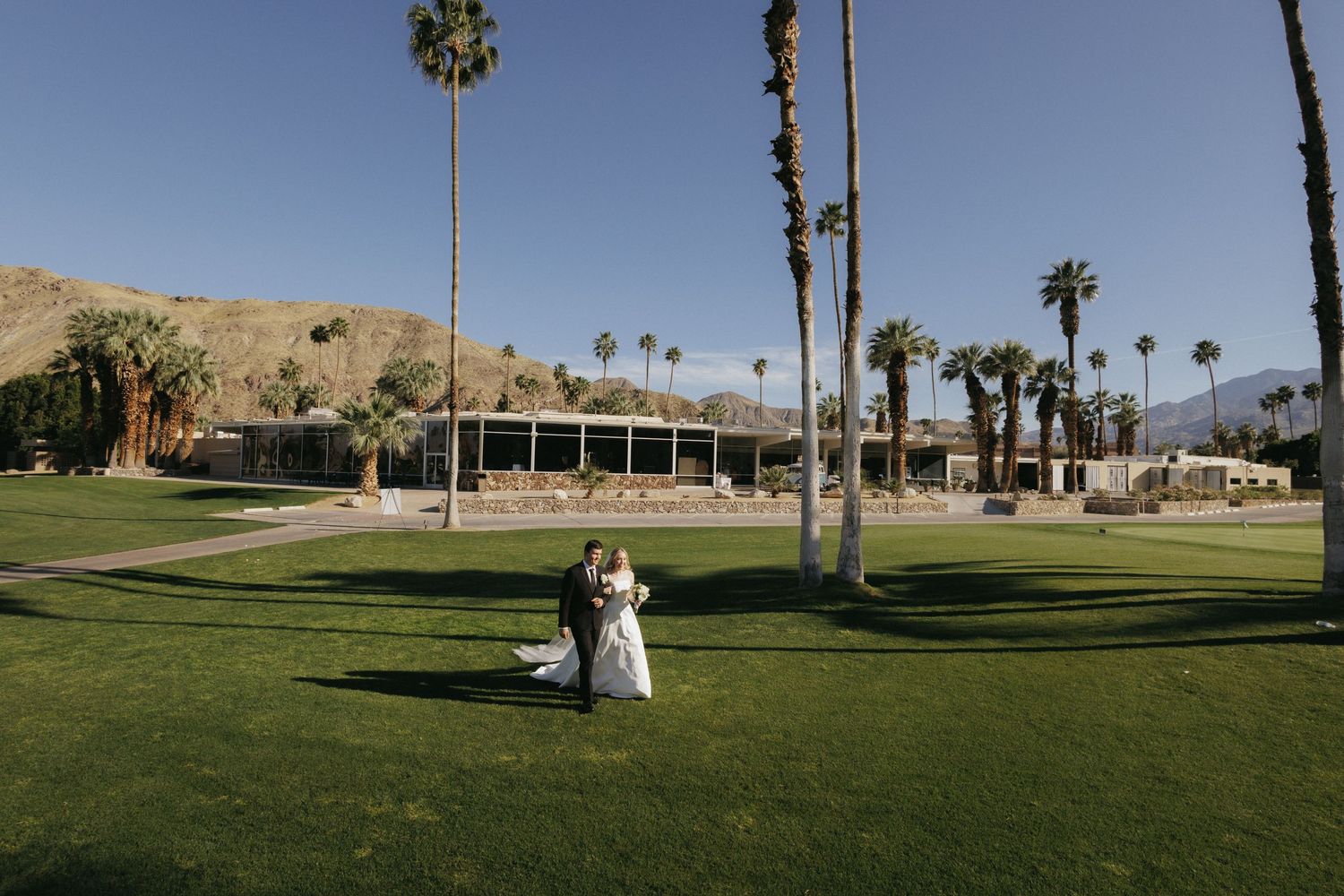 Bride and groom share a romantic moment on a vast green lawn with palm trees and mountain views at Palm Springs venue.