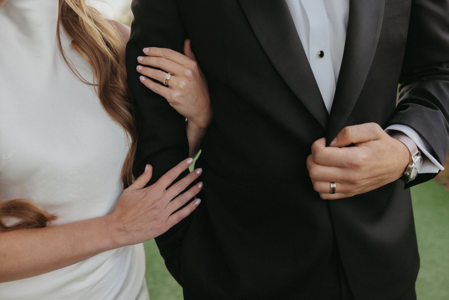 Close-up of bride and groom in wedding attire, showing rings and elegant black tuxedo with white dress.