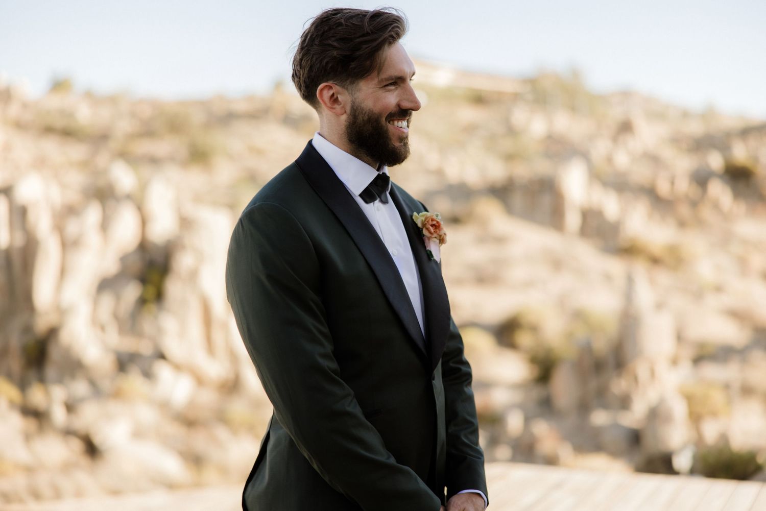 A person in a black suit and tie stands on a rocky mountainous terrain against a bright background.