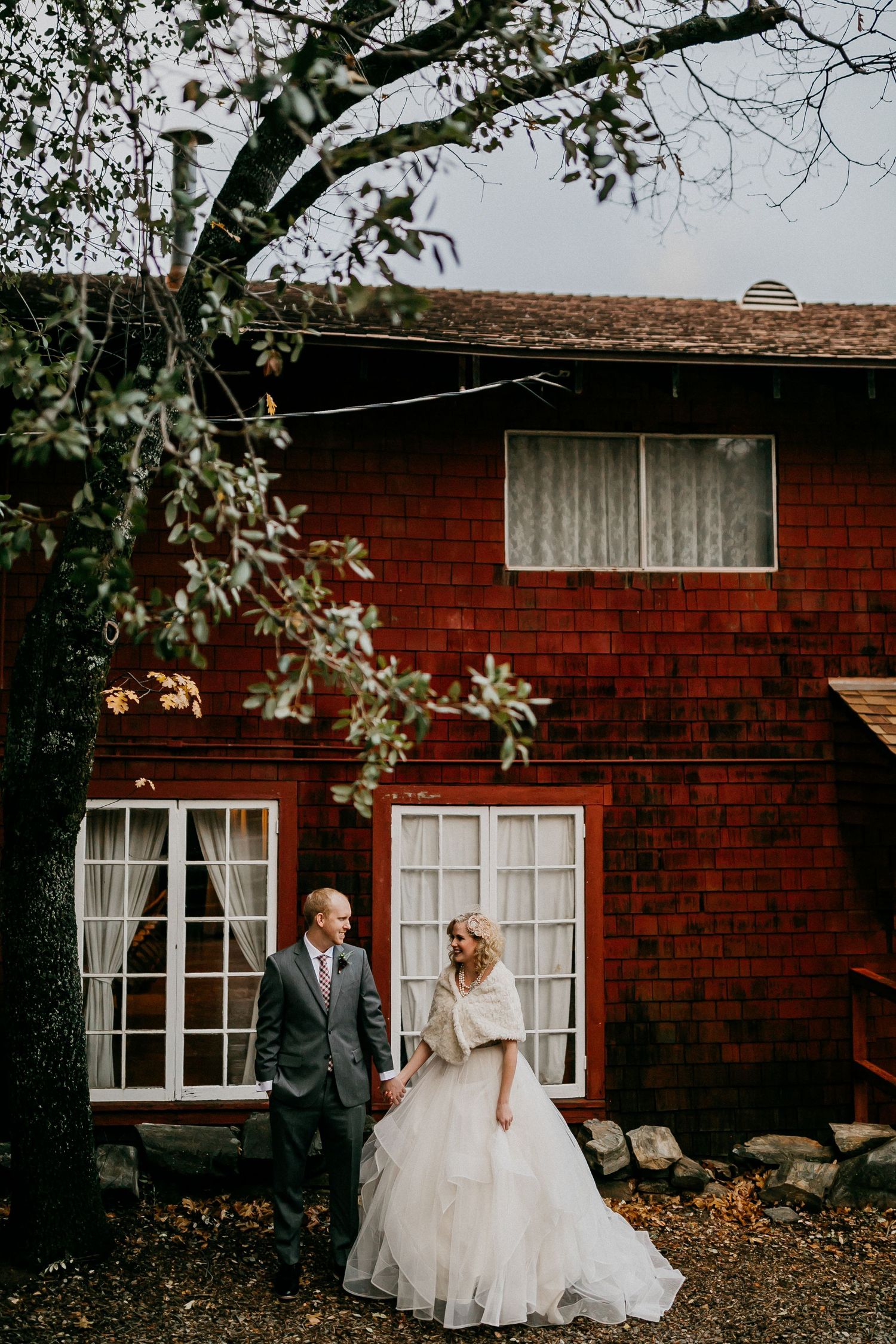 A couple poses together against a red brick building with white windows on their wedding day.
