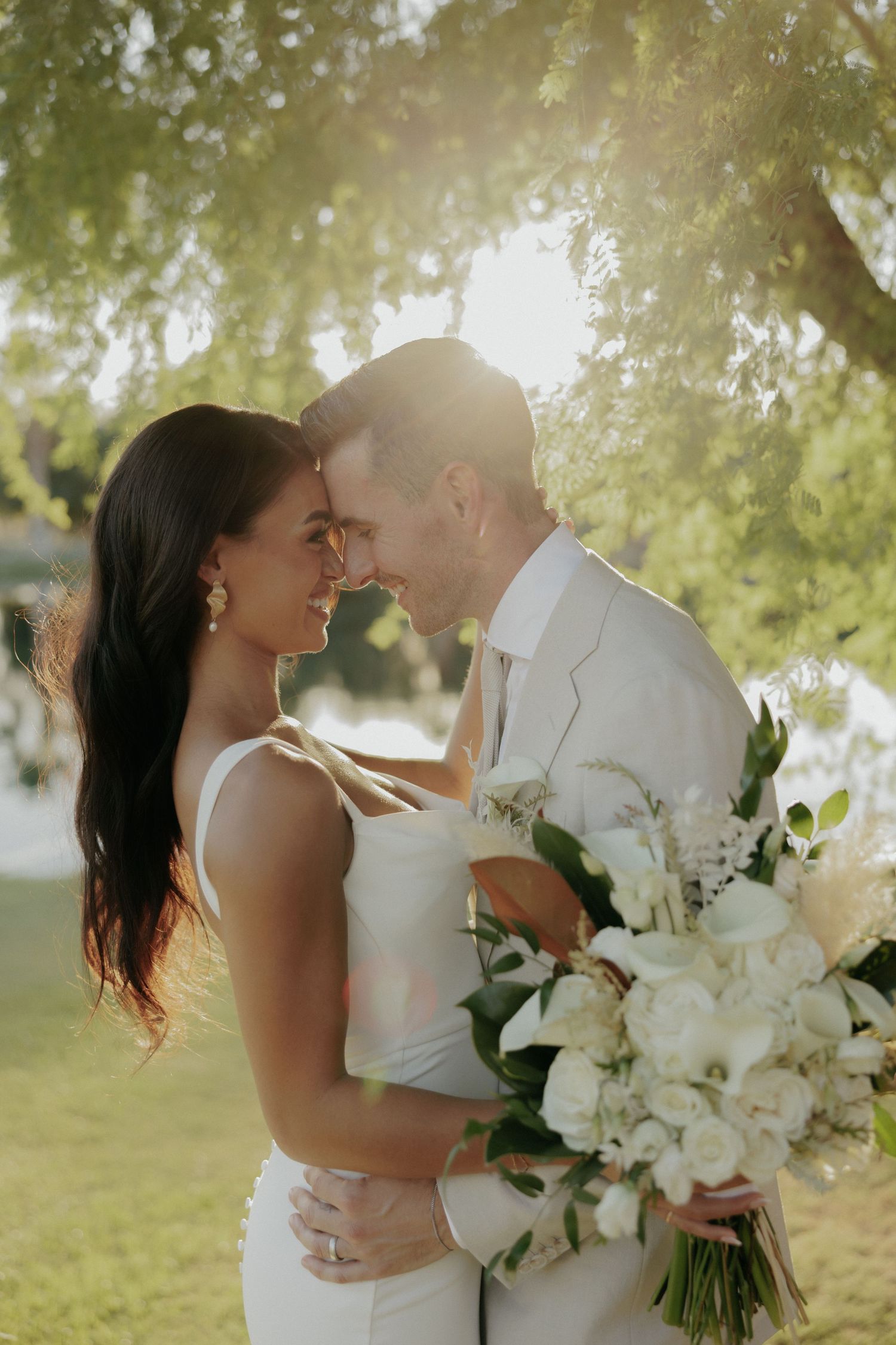 Newlywed couple shares intimate moments outdoors with sunlit trees and white floral bouquet during wedding photoshoot.