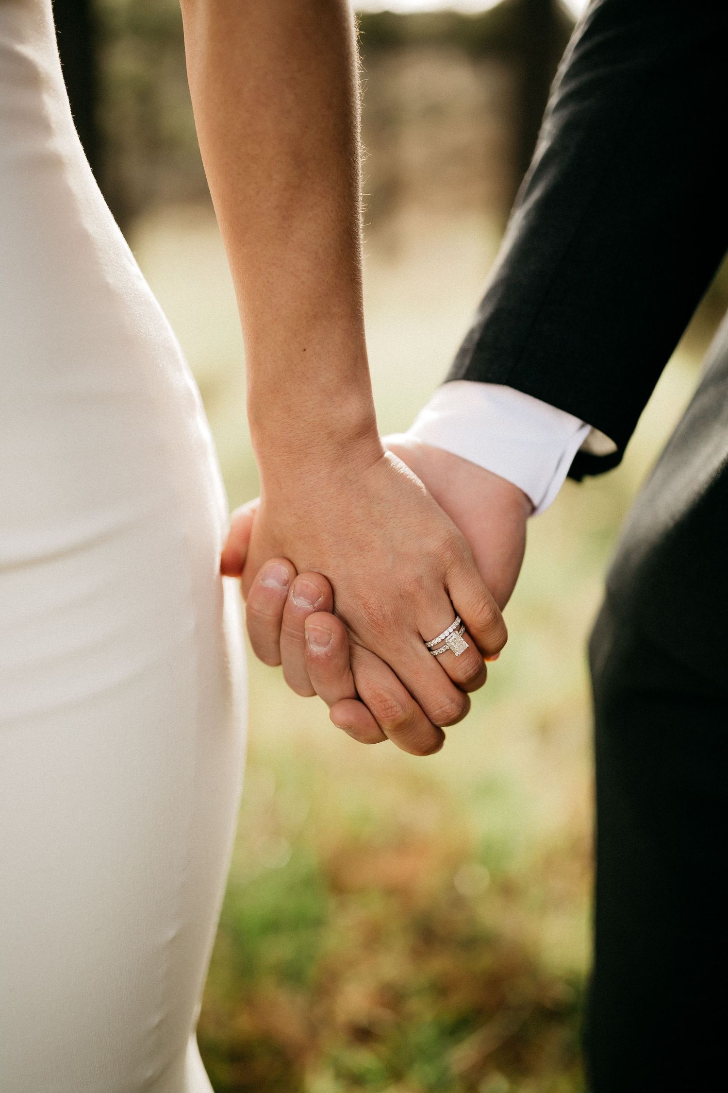 Close-up of newlywed couple holding hands with wedding rings visible against a blurred outdoor background.