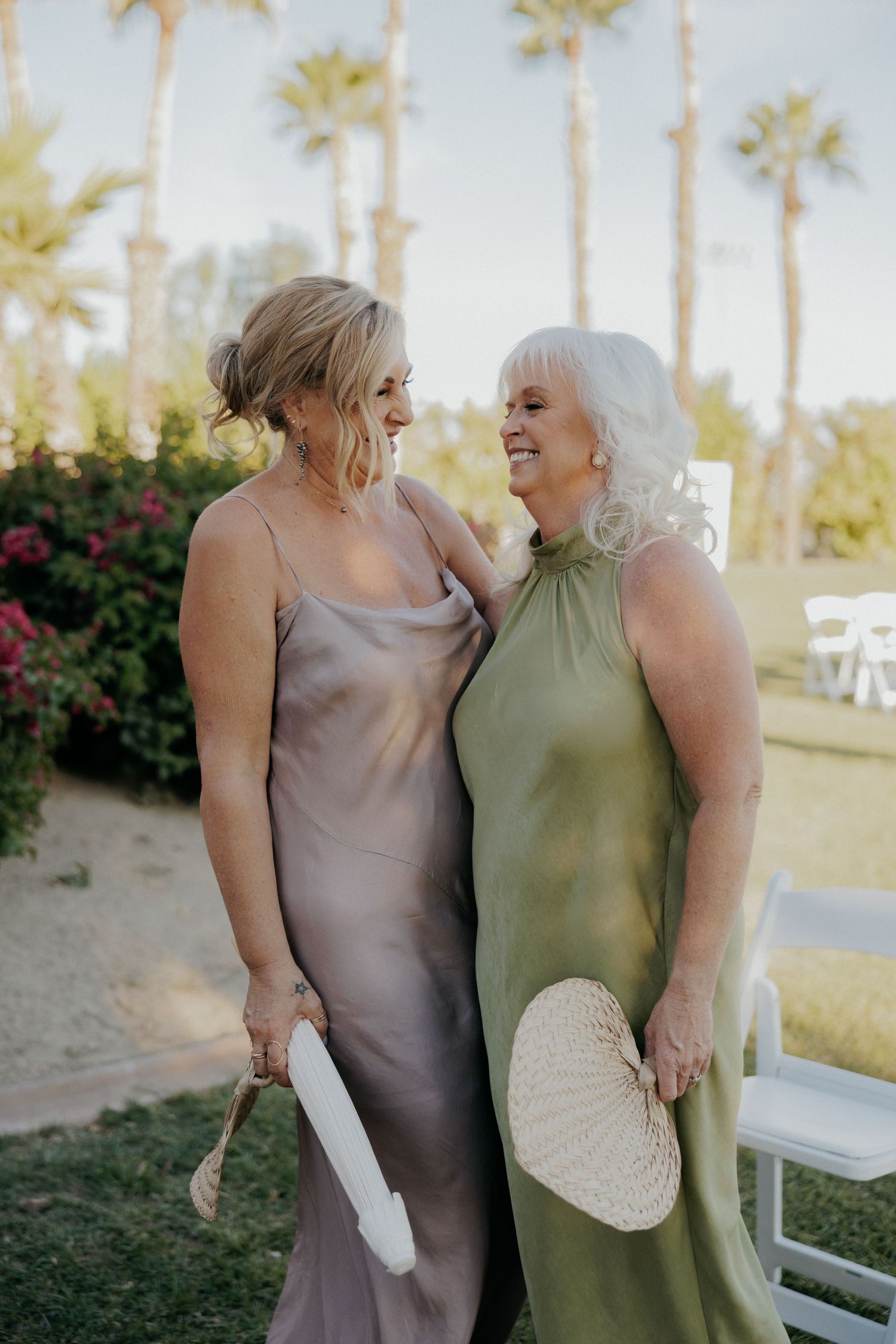 Two women in elegant dresses laugh together while standing outdoors with palm trees and flowers in the background.