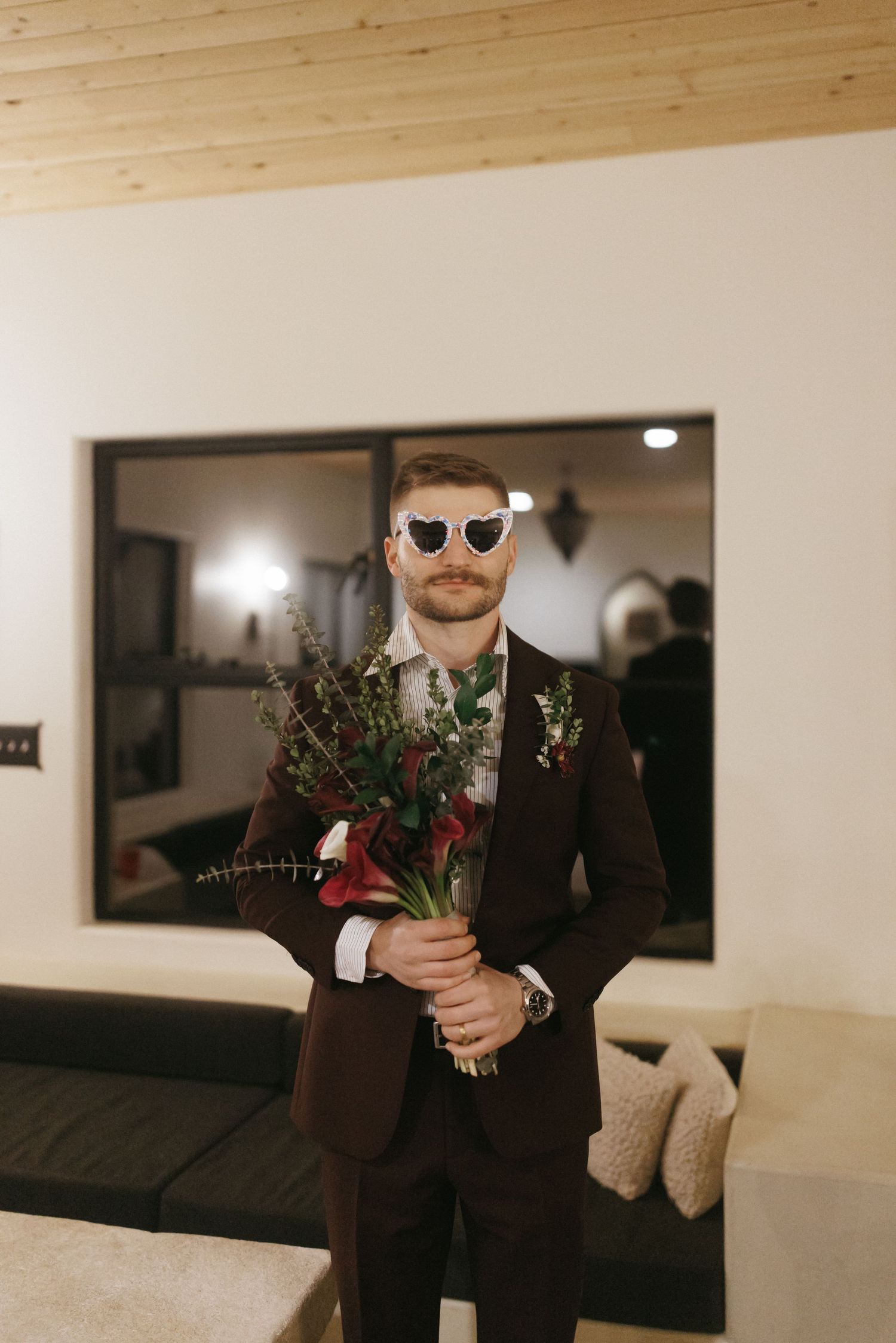 Stylish groom in dark suit and heart sunglasses holds wildflower bridal bouquet indoors before wedding.