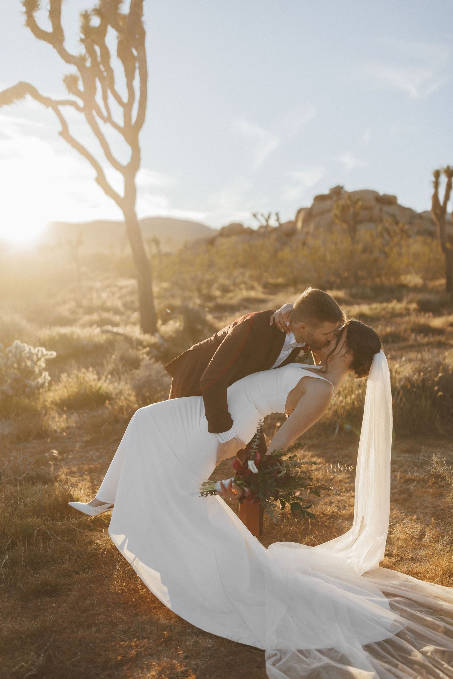 Groom dips bride for a kiss at sunset in Joshua Tree desert with flowing wedding veil.