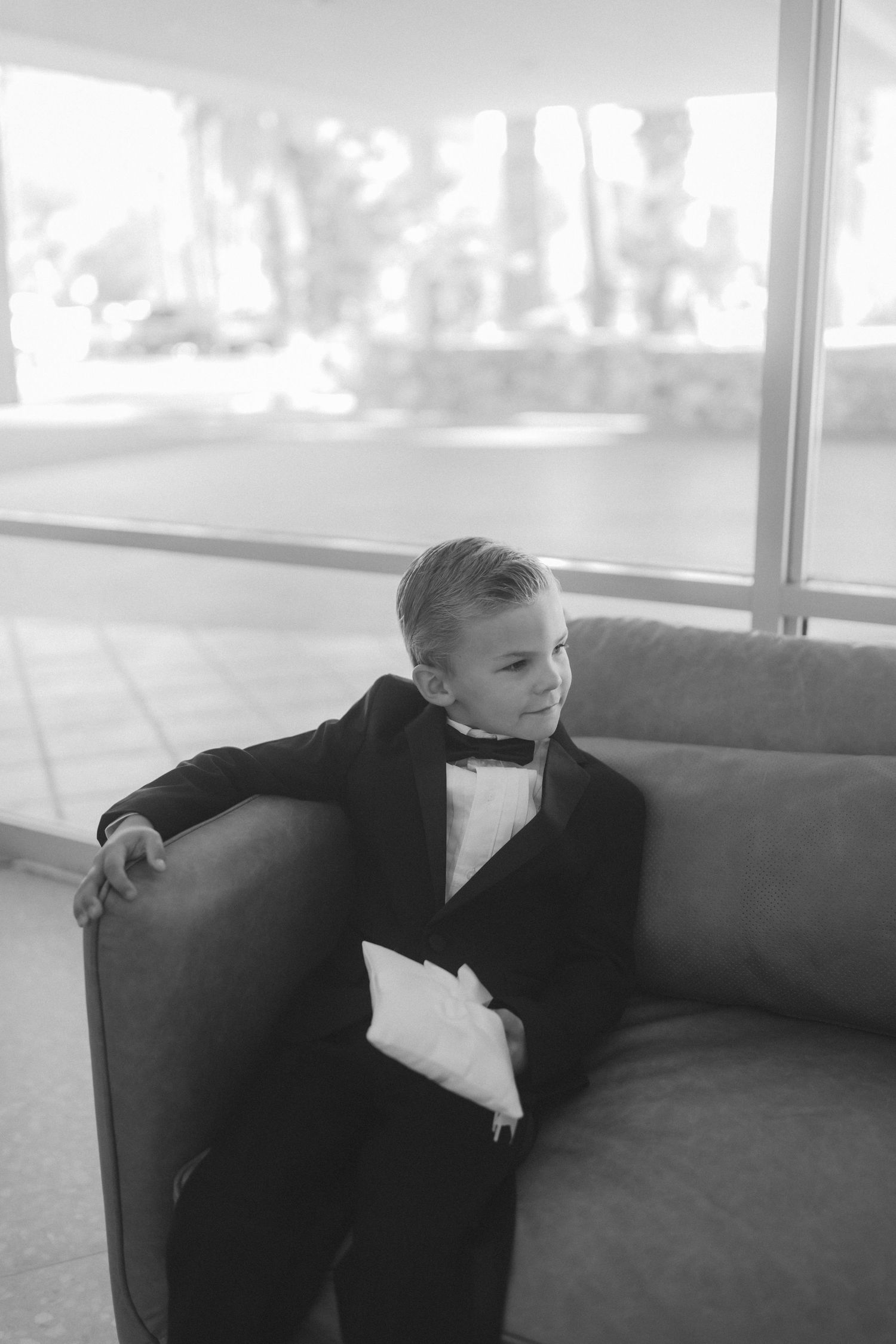 A young boy in a tuxedo with bow tie sits on a gray sofa holding a pillow, serving as ring bearer at a wedding.