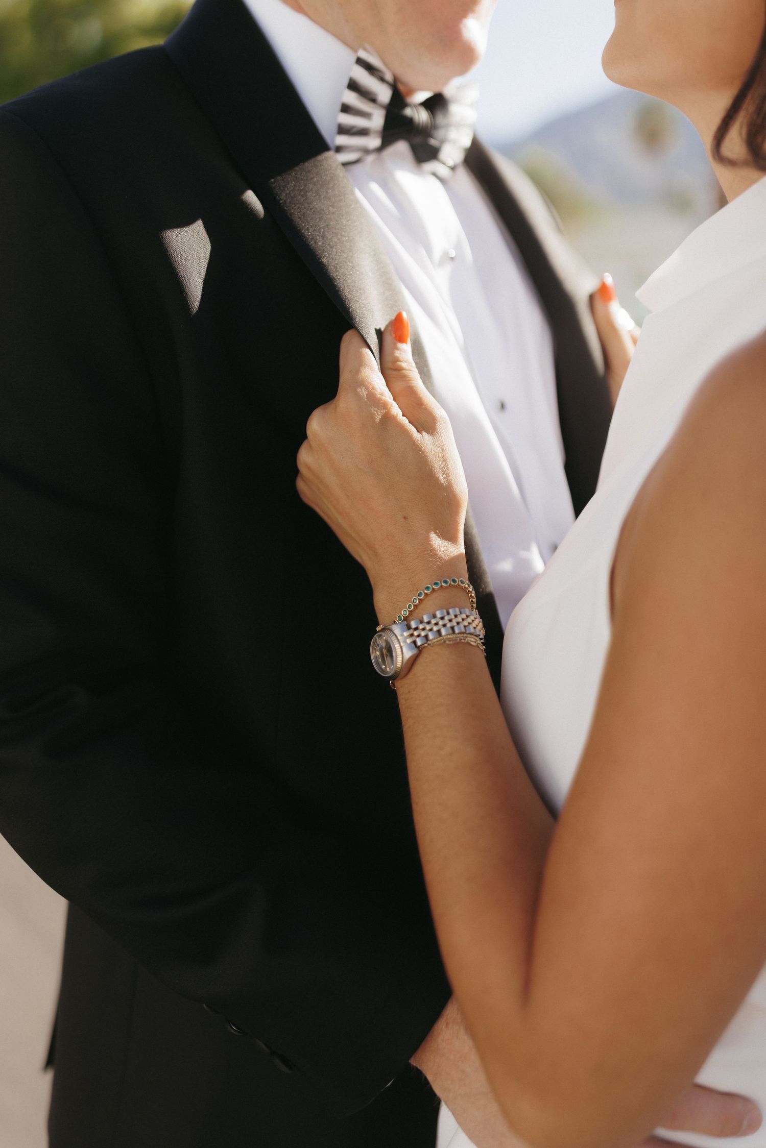 Bride with red nails and silver watch adjusts grooms black tuxedo lapel with striped bow tie outdoors.