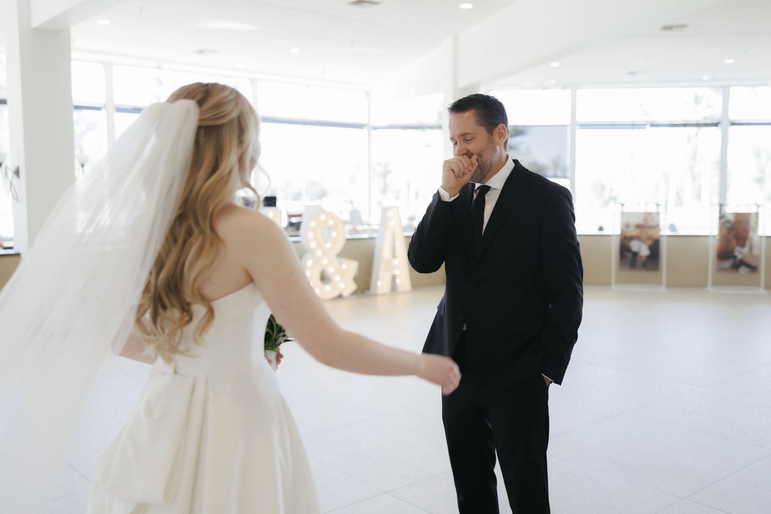 Emotional groom wiping tears as bride in white dress and veil holds his hand during wedding first look.