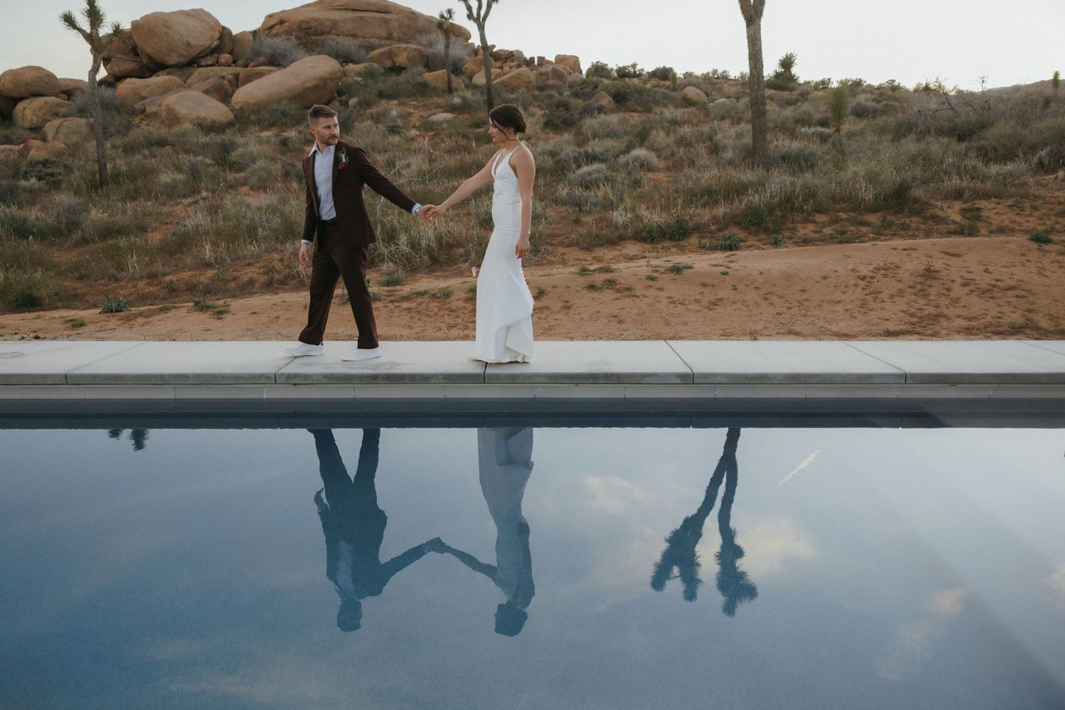 Bride and groom walking hand in hand beside a reflective pool in a stunning desert landscape setting.
