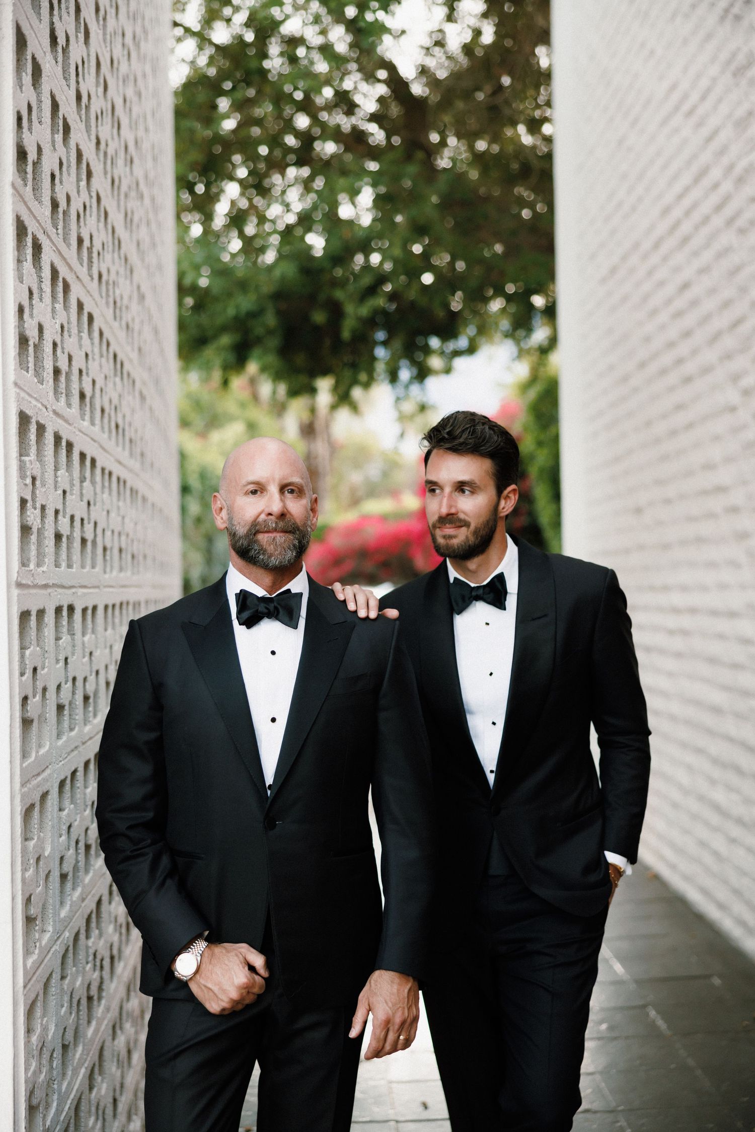 Two men in black tuxedos and bowties share a joyful moment laughing together in a white hallway.