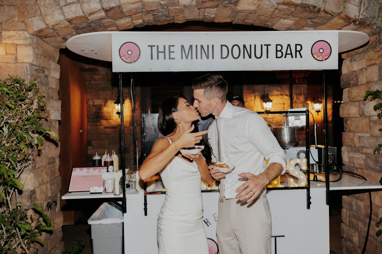 A custom donut station with a decorative sign creates a sweet treat experience at an elegant indoor wedding reception.