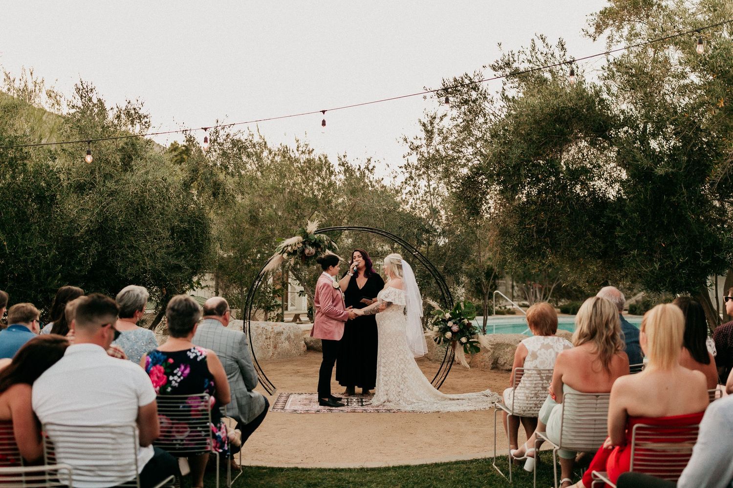 An outdoor wedding ceremony under string lights and trees with guests seated on wooden chairs.