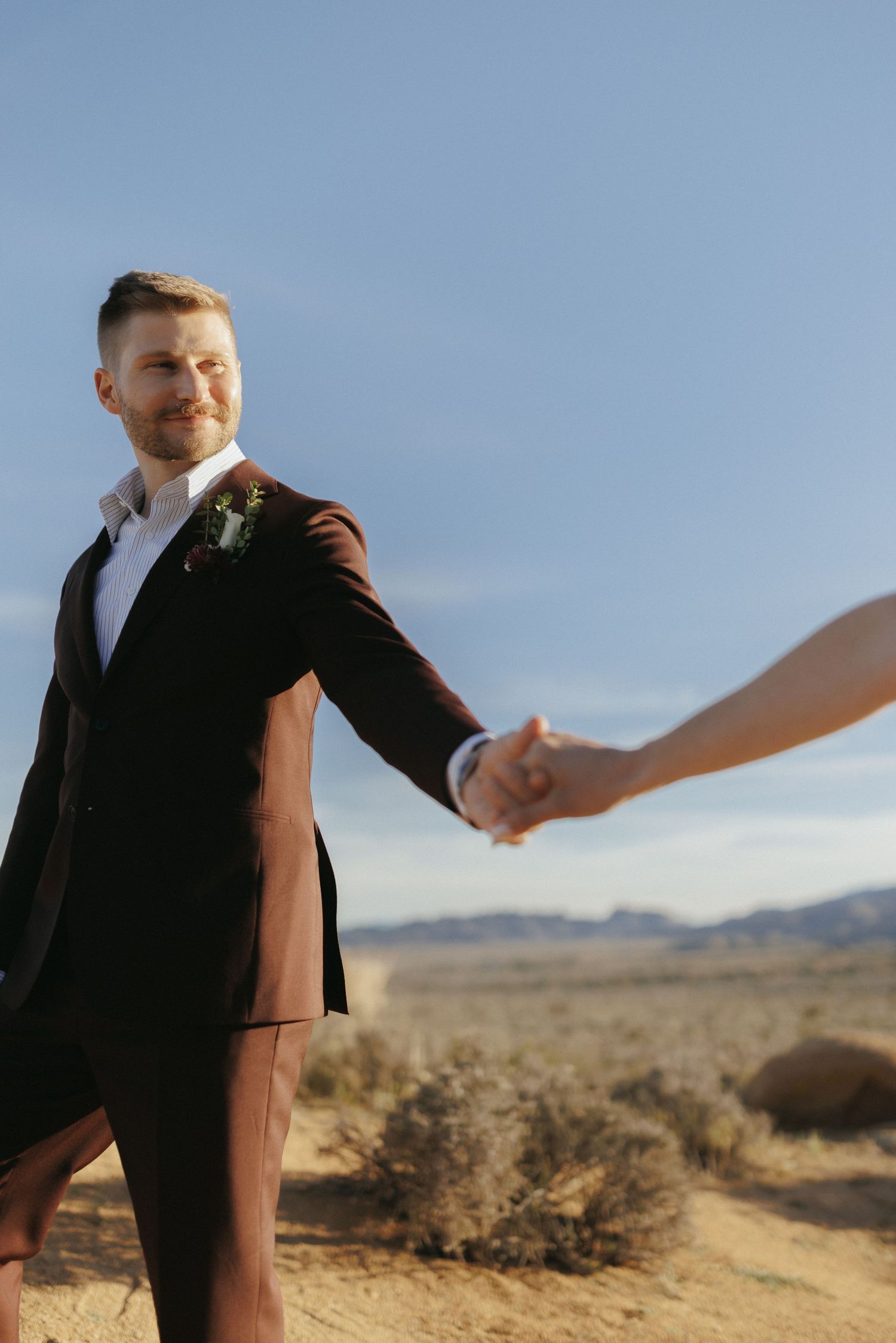 Groom in dark suit with boutonniere holds bride's hand in sunlit desert landscape.
