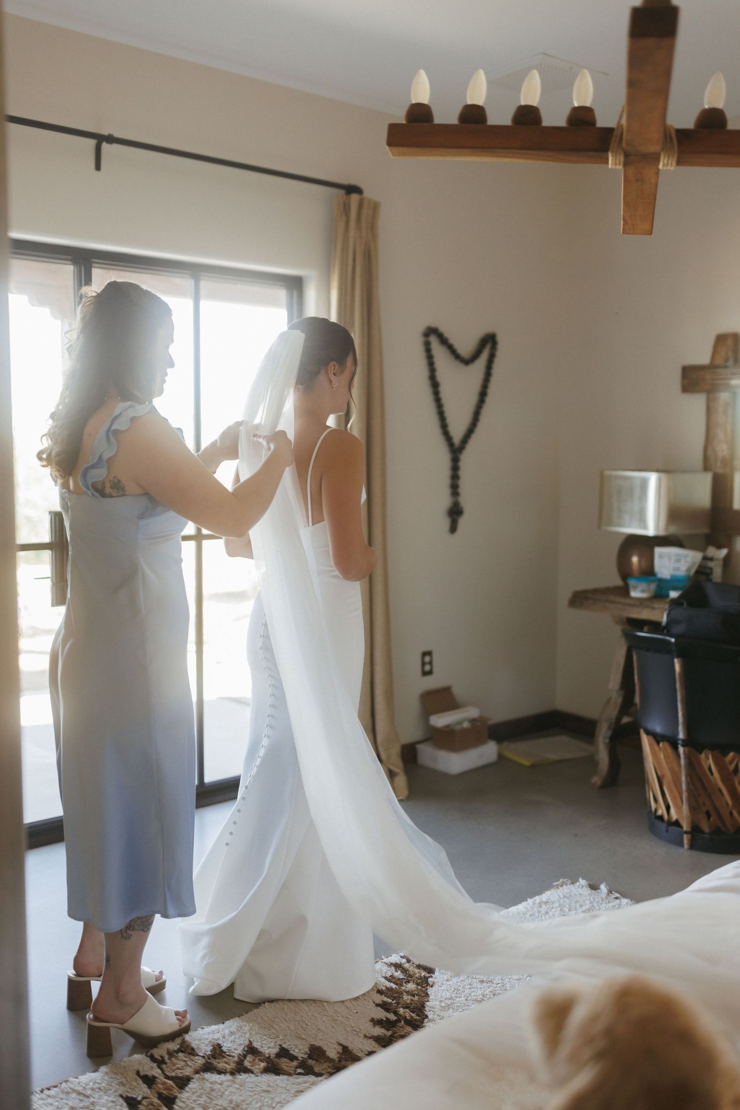 Bridesmaid helping bride button her wedding dress in a warmly lit rustic bedroom before the ceremony.