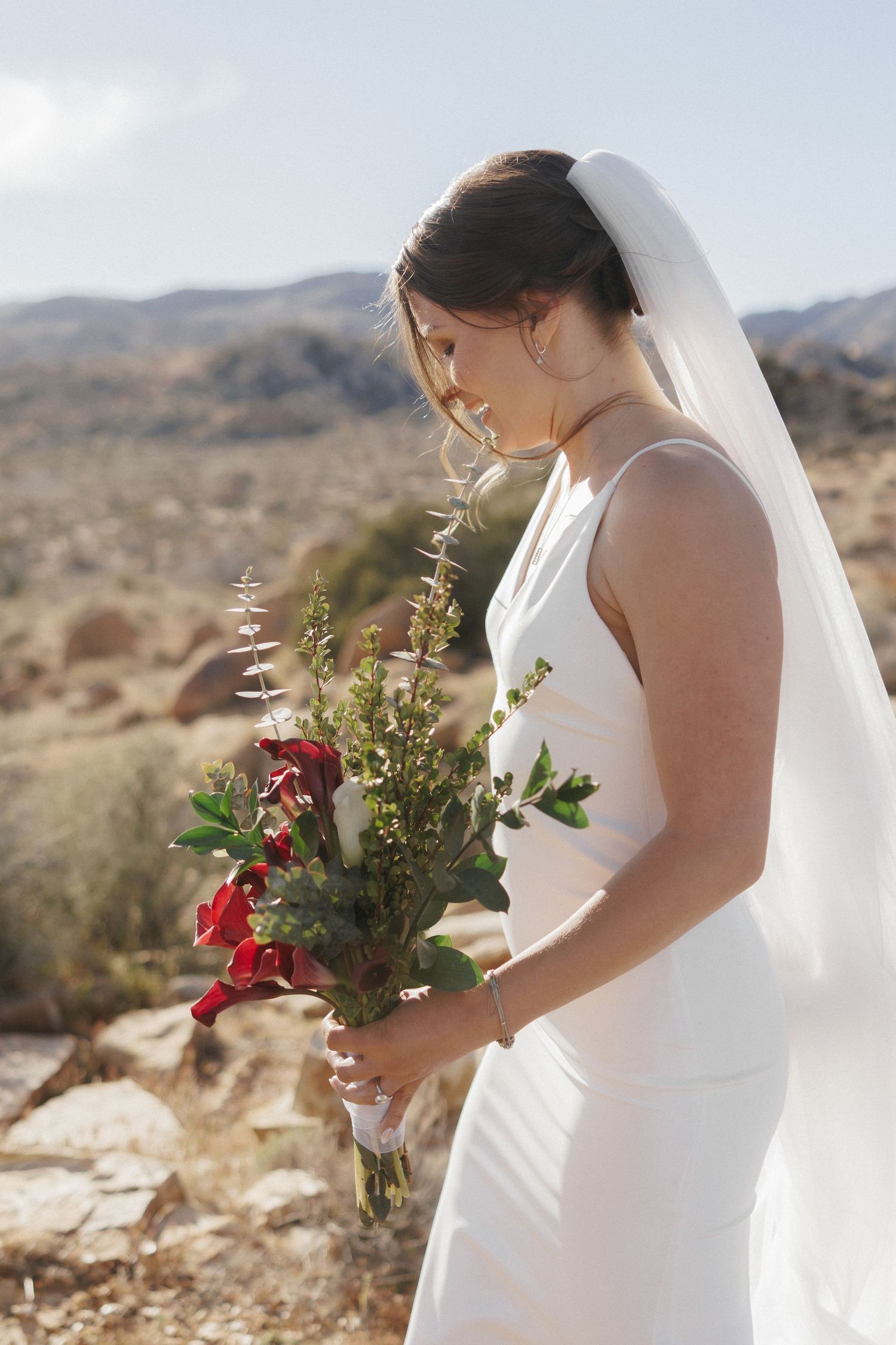 Bride in white gown holding red and green bouquet with desert mountains behind her.