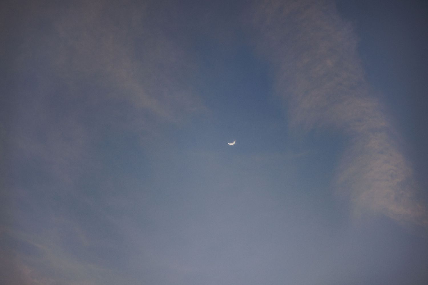Crescent moon visible against soft blue sky with wispy clouds at dusk.