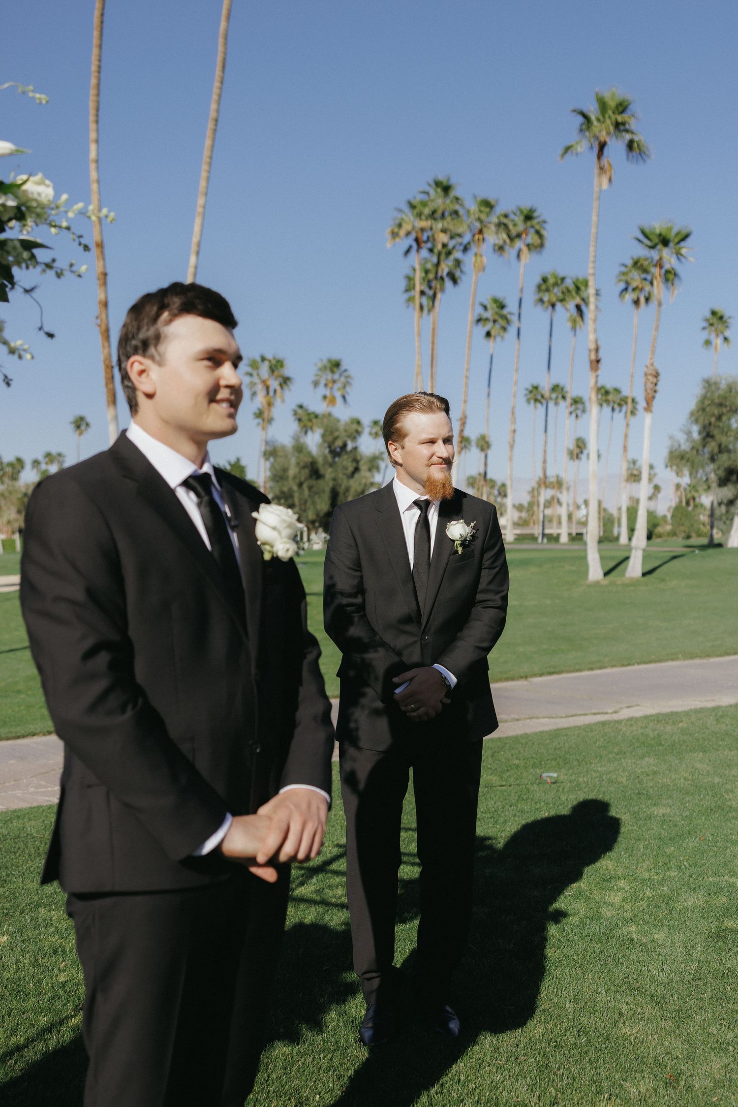 Two groomsmen in black suits with white boutonnieres standing on green lawn with tall palm trees.