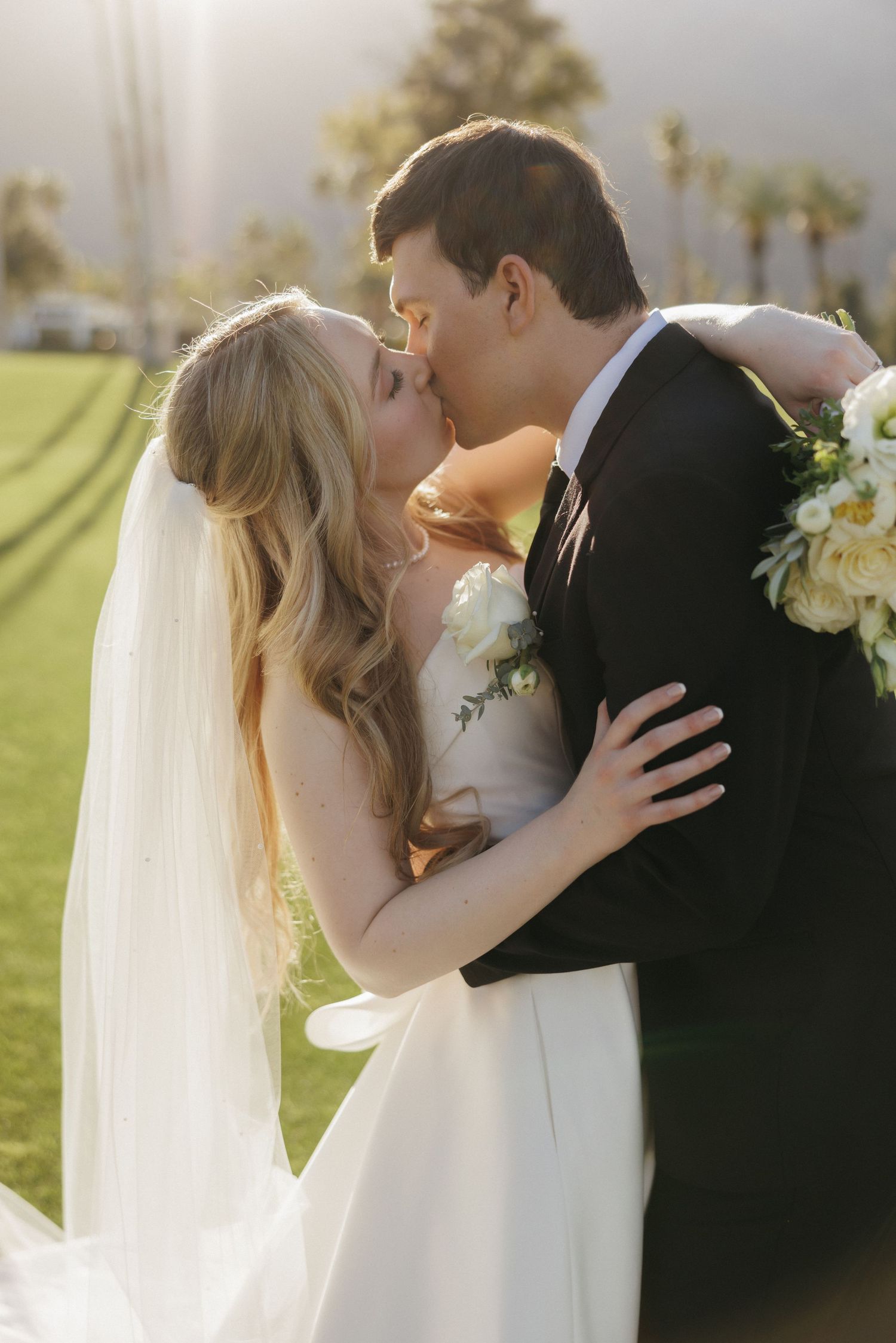 Bride and groom share romantic kiss on sunlit green lawn with palm trees in background.