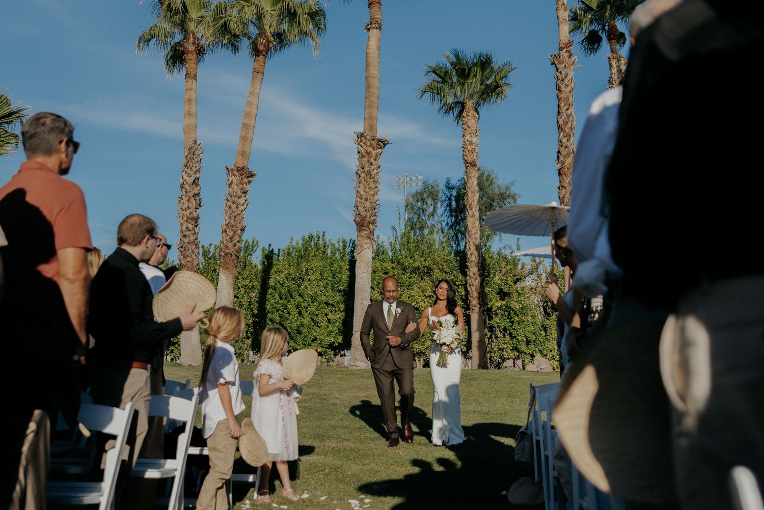 Wedding guests gather beneath tall palm trees on a grassy lawn during an outdoor ceremony.