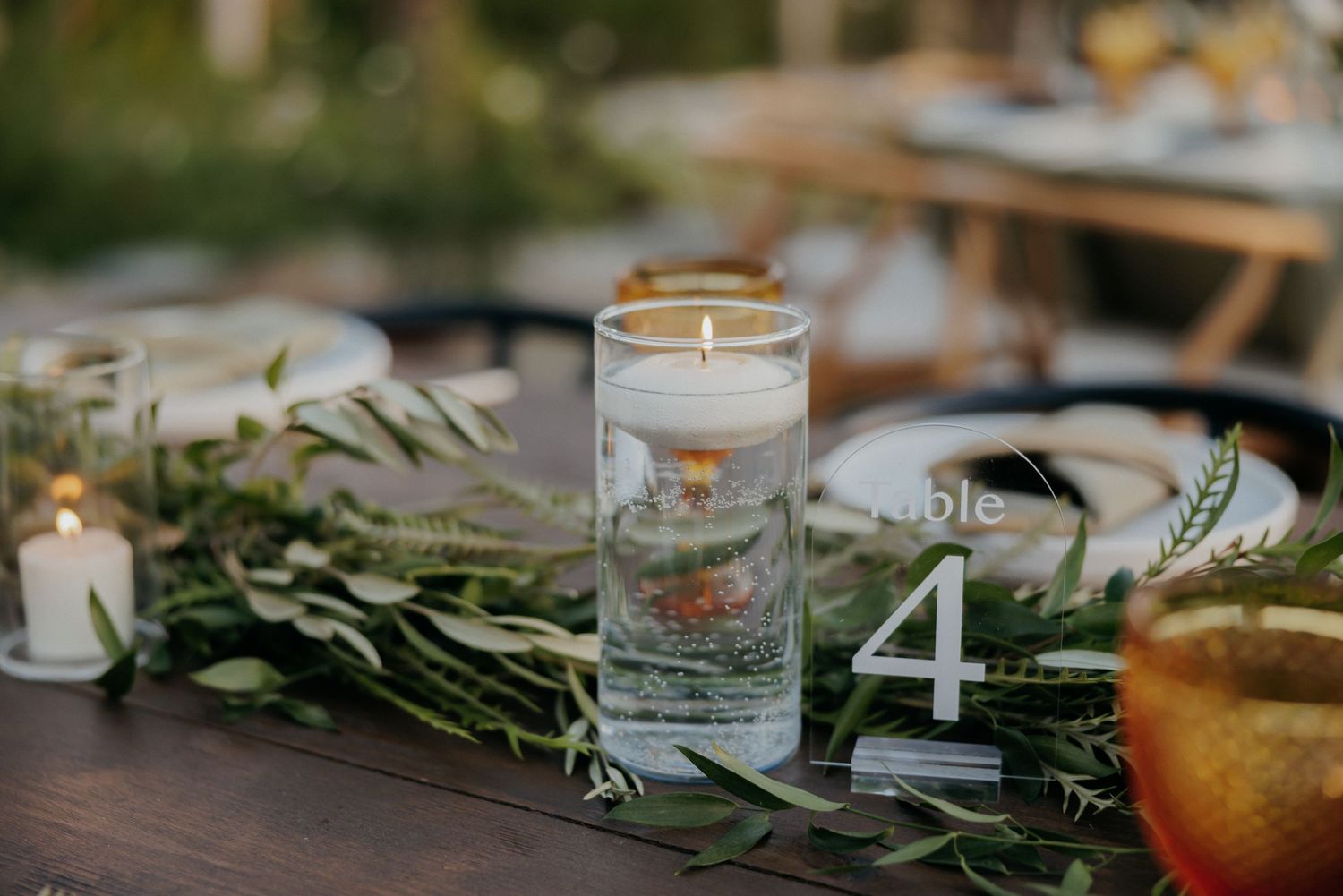 Rustic table setting with frosted glass candle holders and fresh greenery garland against warm wooden surface.