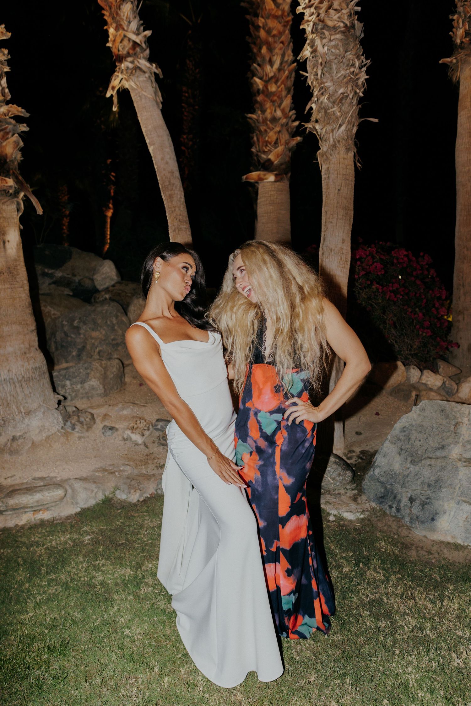 Dancing outdoors at night, two people in contrasting dresses move together under palm trees in a garden setting.