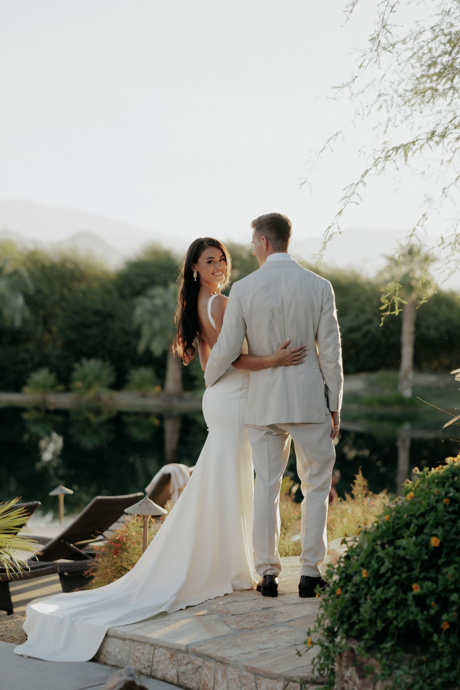 A couple shares a romantic moment on a stone wall at sunset, the bride's white dress flowing in the golden light.