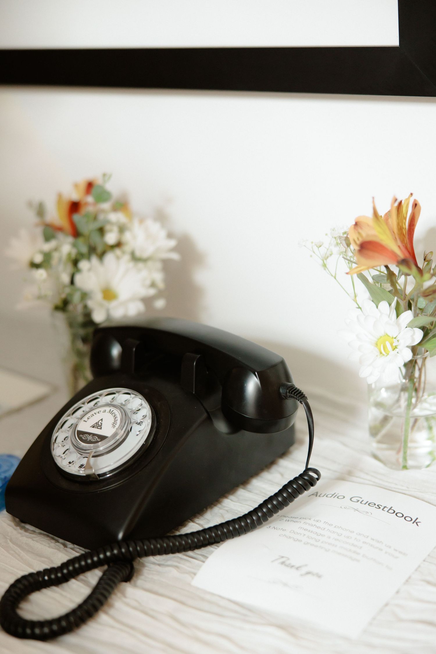 Vintage black rotary telephone sits on white surface next to small vase of flowers in artistic composition.