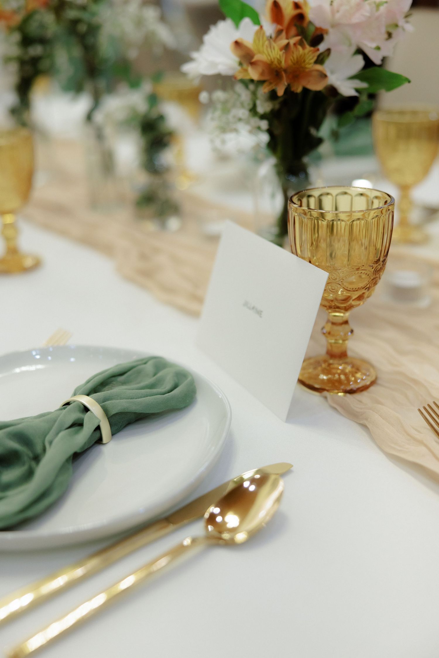 Elegant place setting with gold flatware, mint green napkin, and amber goblet on white linen tablecloth.