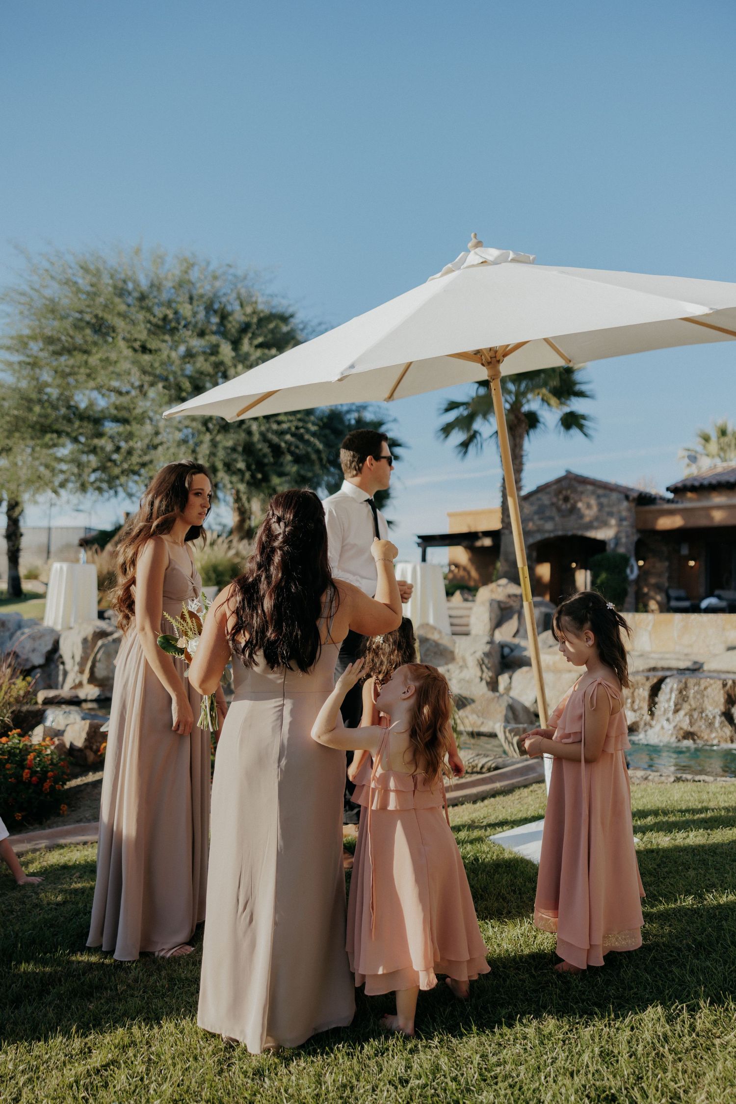 Bridesmaids in blush pink dresses hold white parasols while standing on a sunlit lawn with palm trees in the background.