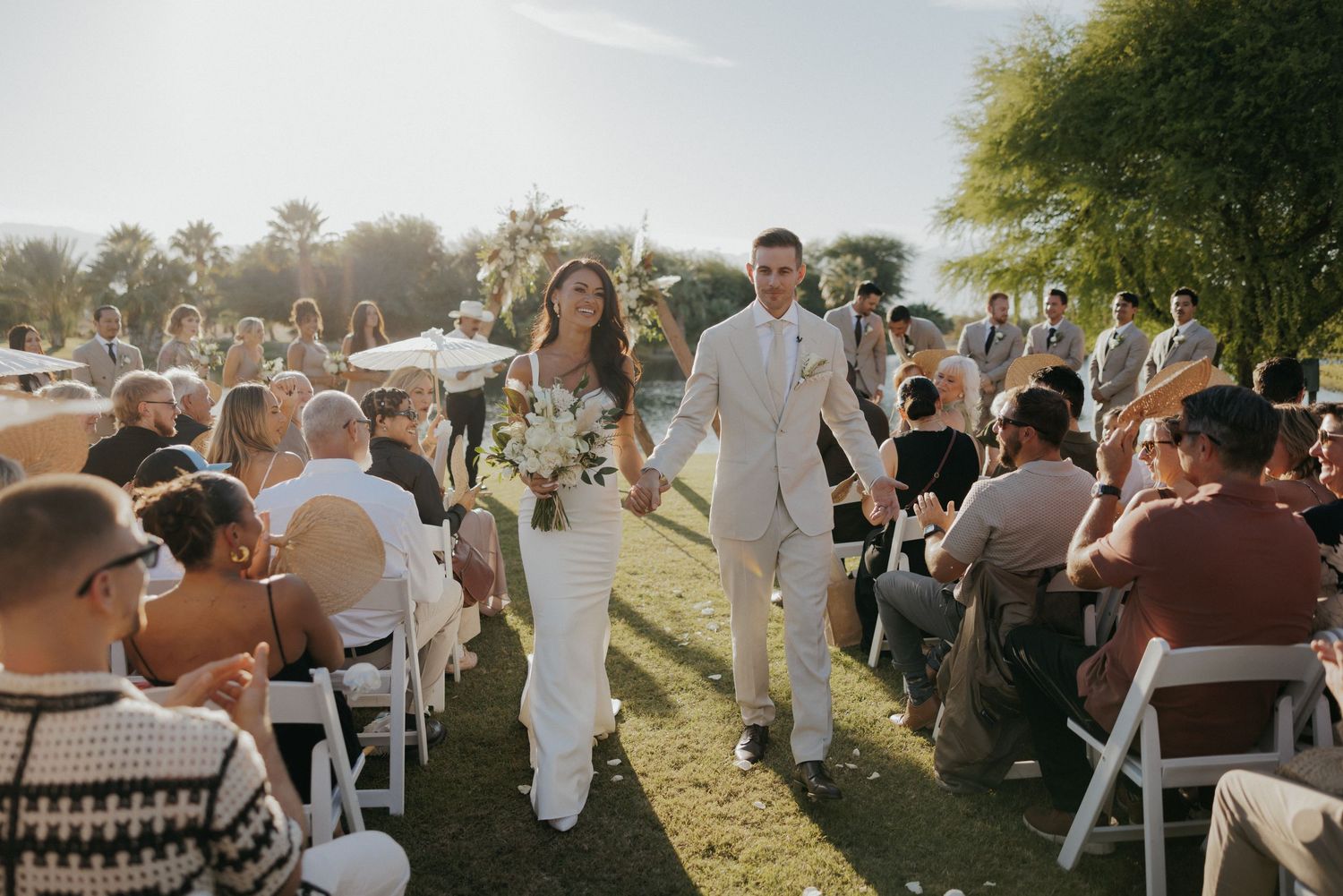 Wedding guests enjoy a sunny backyard celebration while seated on white chairs at an outdoor reception.