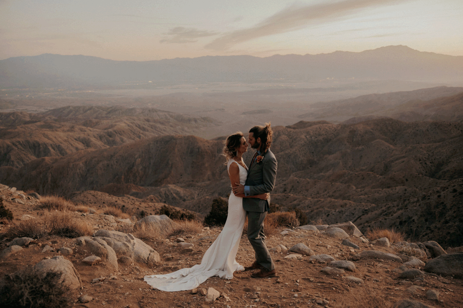 Couple sits on edge of rugged mountain peak overlooking vast desert landscape during golden sunset hour.
