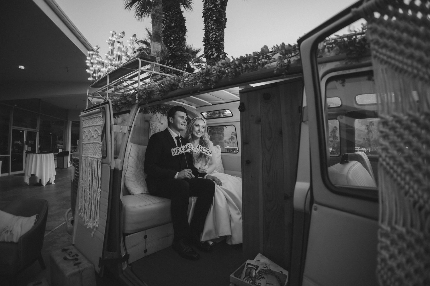 Bride and groom sit together inside a decorated vintage van photobooth at an outdoor evening wedding reception.