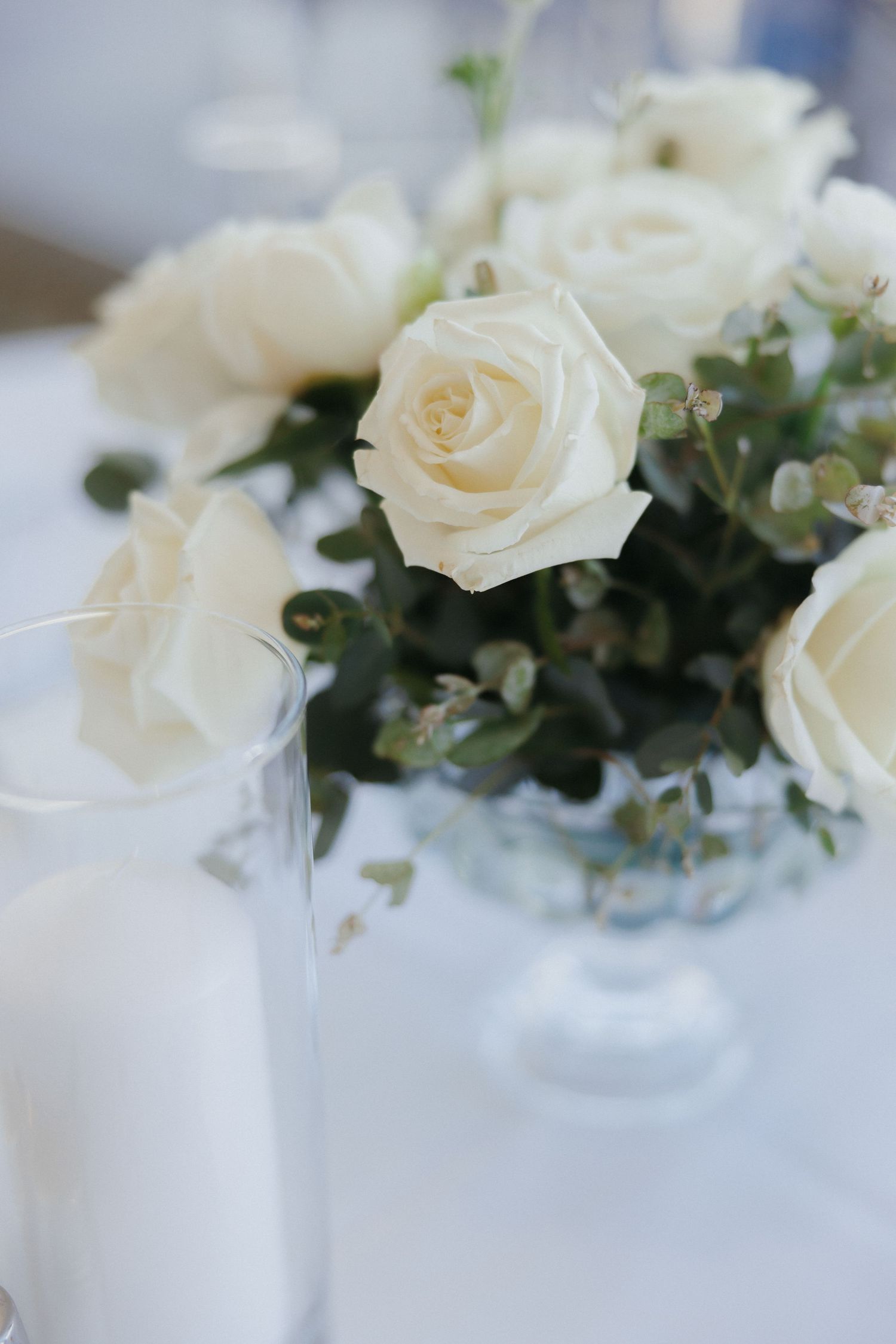 White roses and eucalyptus centerpiece in crystal vase on white linen wedding reception table.