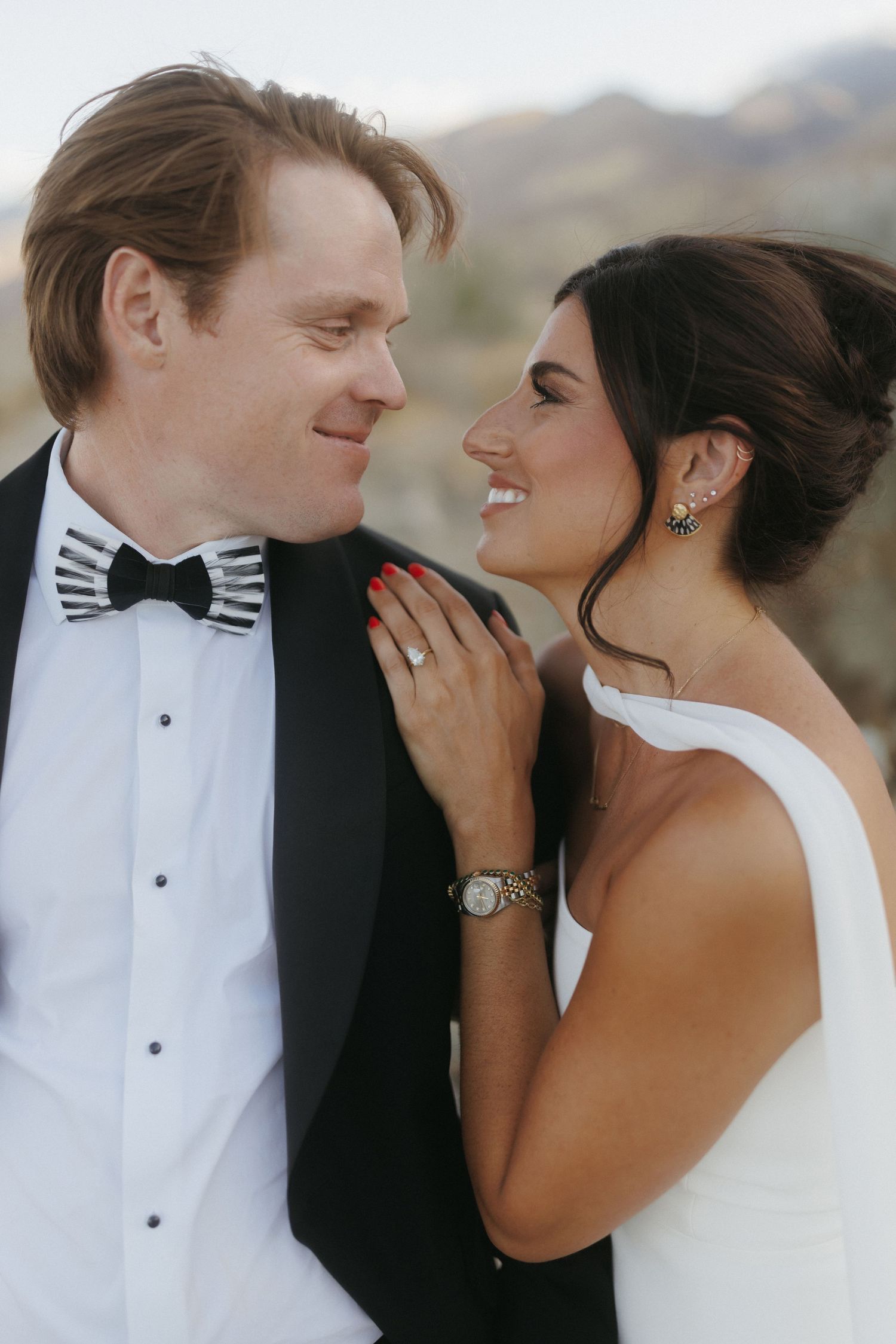 Smiling bride and groom gaze at each other, bride wearing diamond ring, gold watch, and floral earrings.