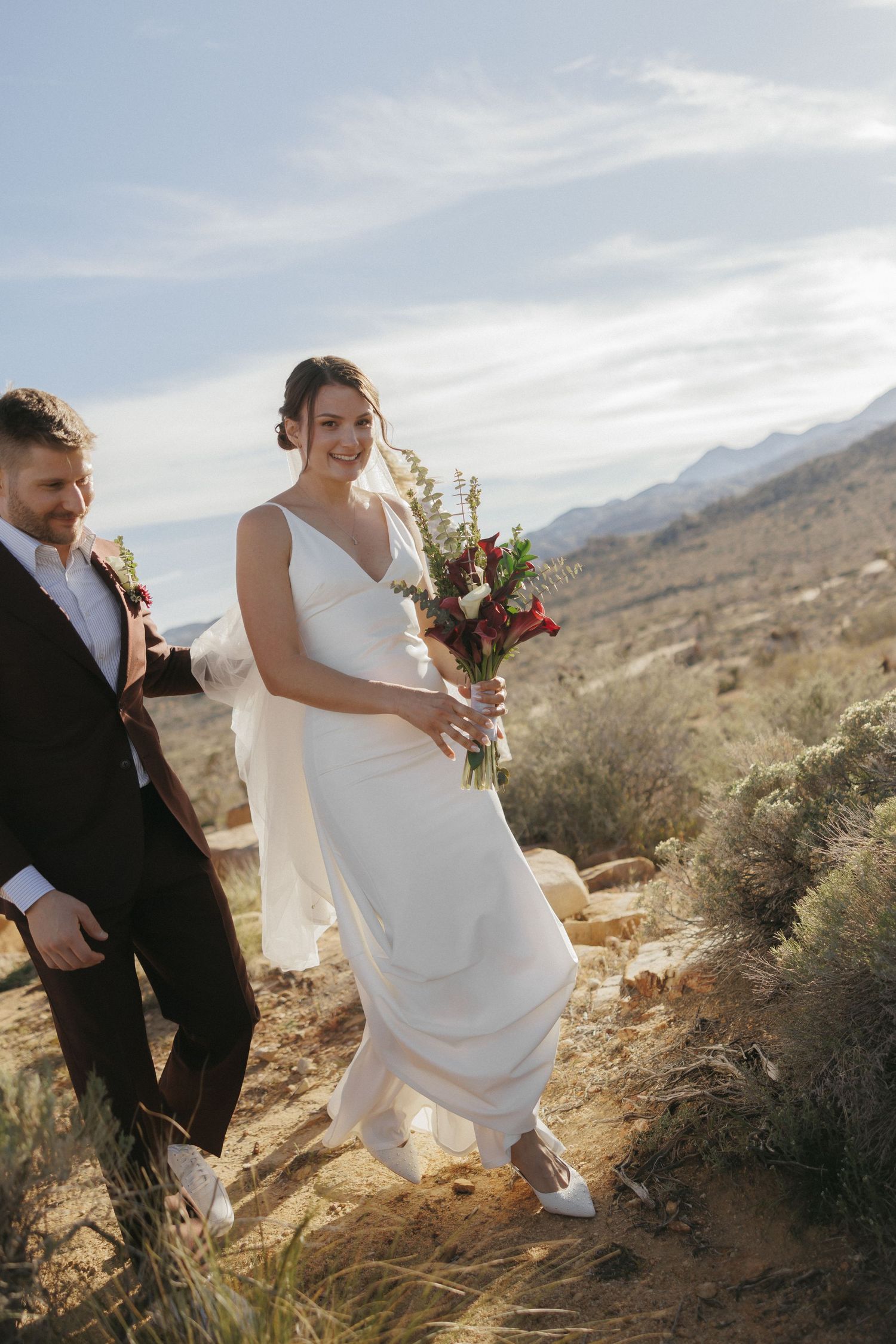 Bride and groom walking through desert landscape, bride holding red bouquet in white gown, mountains in background.
