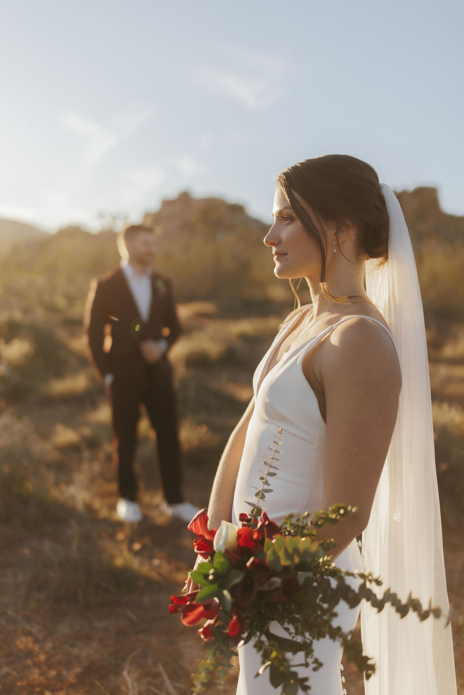 A bride in a white gown holds red bouquet with groom standing in desert background.
