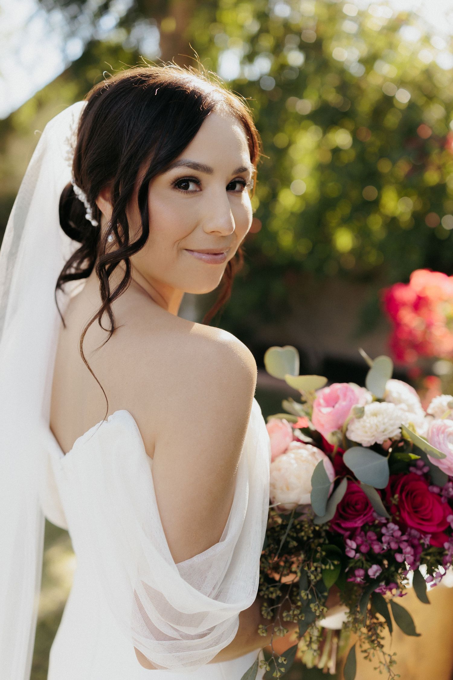 A bride in a white wedding dress with draped cowl back detail turns to smile while holding a vibrant floral bouquet.