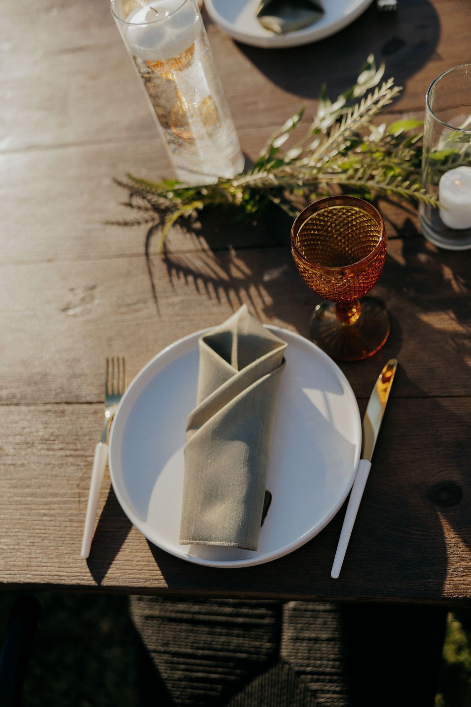 Place setting on rustic wooden table with white plates, cutlery and glassware in golden evening light.