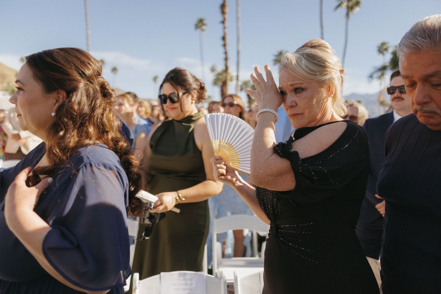 Wedding guest in black dress fans herself with a hand fan at a sunny outdoor ceremony in Palm Springs.
