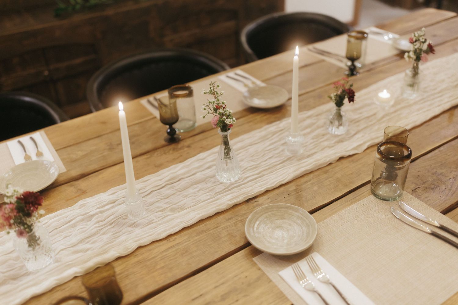 Rustic wooden wedding reception table set with white taper candles, bud vases, and linen table runner.