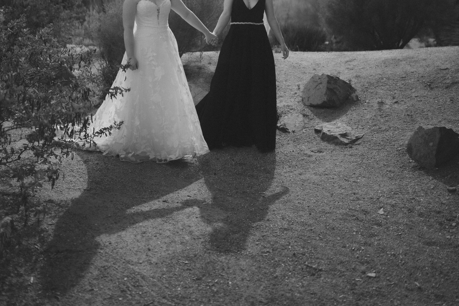 Black and white photo of two women in formal gowns holding hands on a sandy path outdoors.