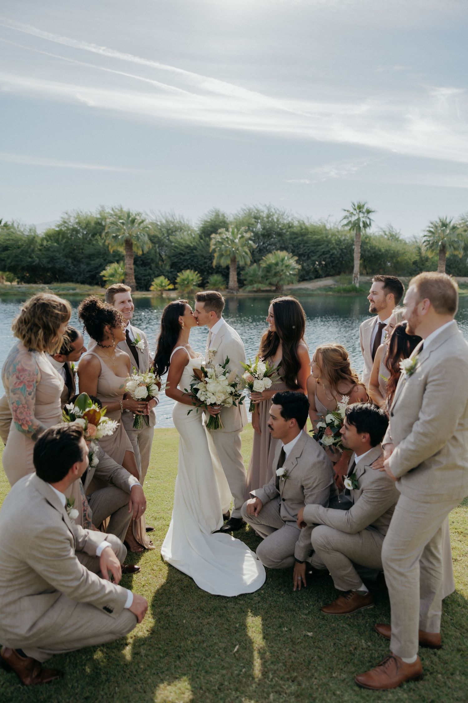 Wedding party poses playfully while sitting on grass by a lakefront during outdoor celebration.