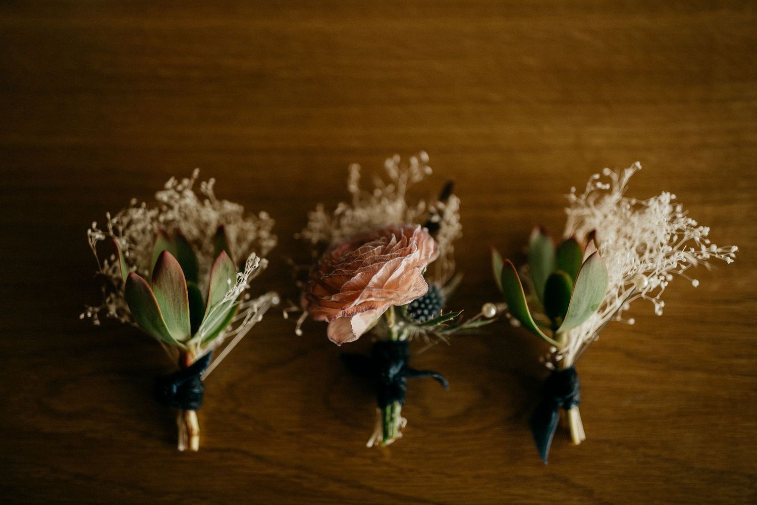 Wedding boutonnieres with delicate flowers lined up in a row on a dark wooden surface.