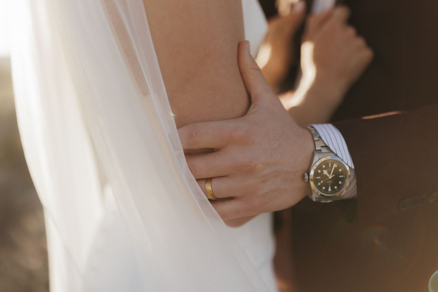 A groom in a suit holds his bride close, wearing a silver watch and gold wedding band.