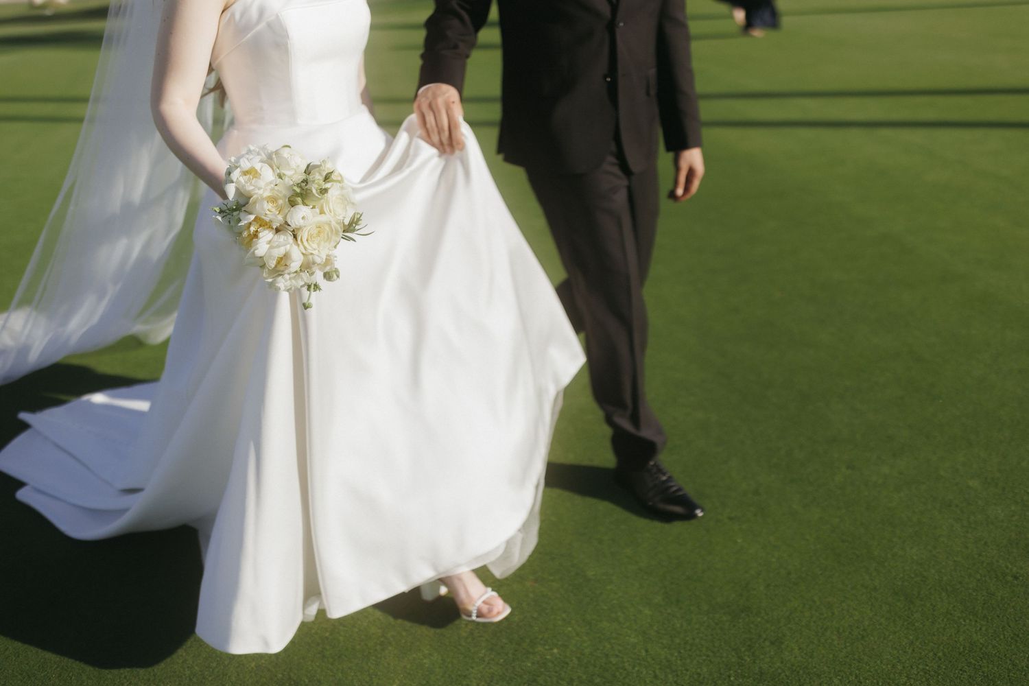 Groom lifts bride's white wedding gown as couple walks across green golf course.