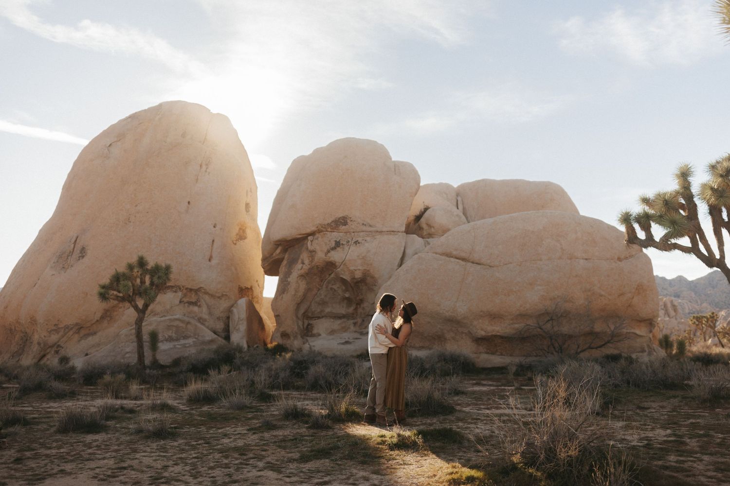 Couple embracing at golden hour among large boulders and Joshua trees in Joshua Tree National Park desert landscape.
