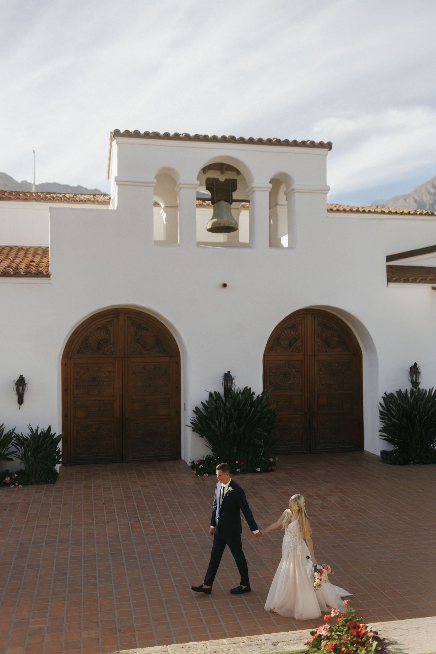Bride and groom holding hands walking in front of a white Spanish-style chapel with bell tower and arched wooden doors.