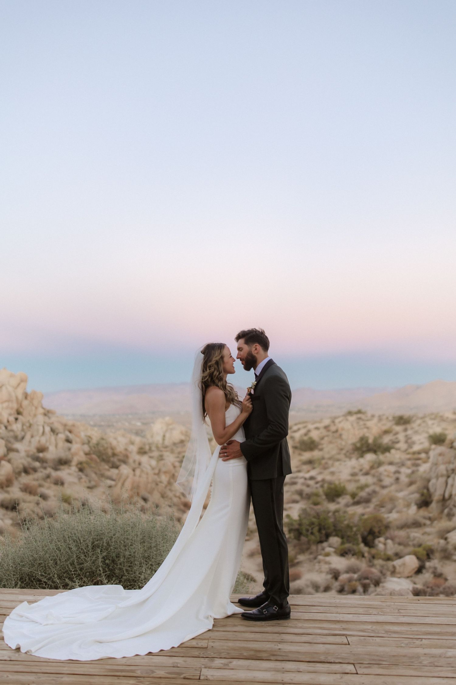 A romantic wedding couple embraces on a wooden deck overlooking a desert landscape at sunset.